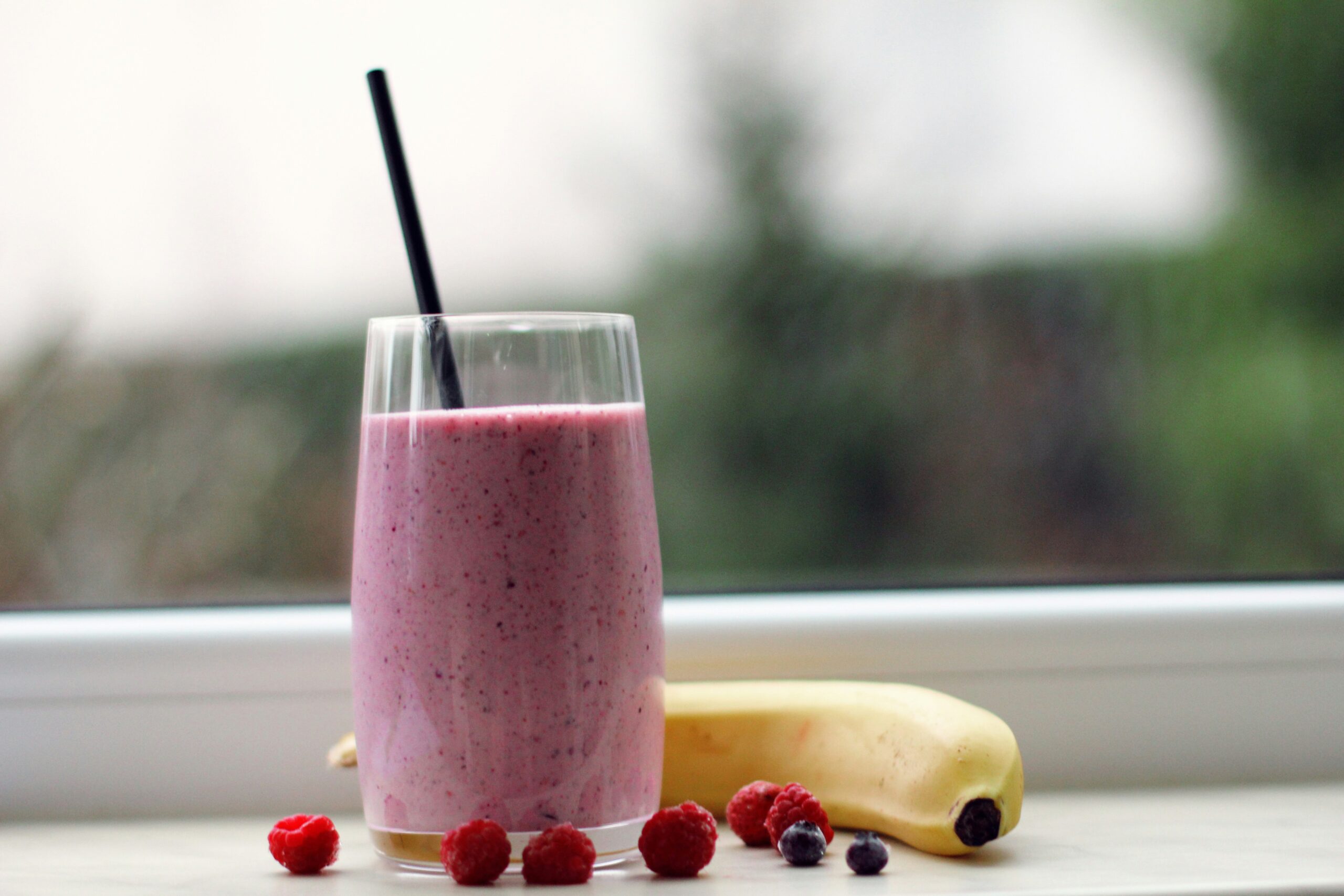 A refreshing berry smoothie in a glass with a black straw, accompanied by fresh raspberries and a banana on a wooden surface, with a blurred outdoor background.