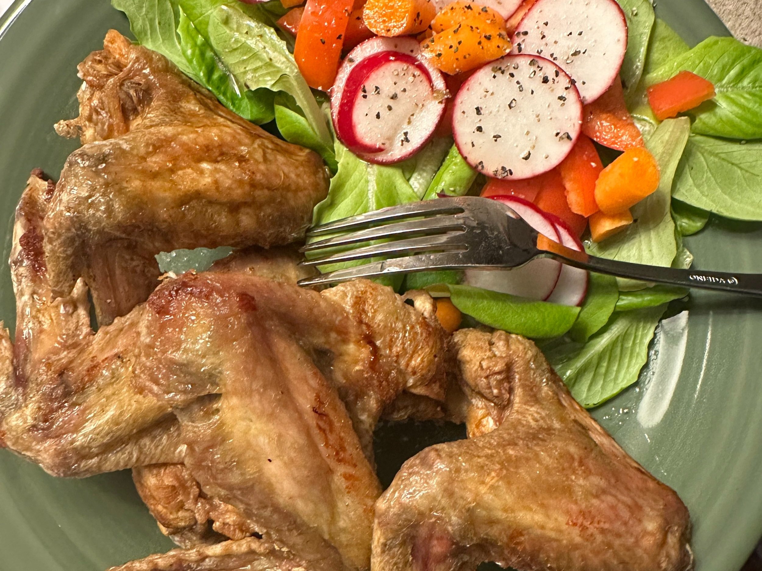 Barb+Peace+-+IMG_4998 Crispy chicken wings served on a green plate alongside a fresh salad featuring sliced radishes, mixed greens, and colorful bell peppers. A fork rests on the plate, highlighting the delicious meal.