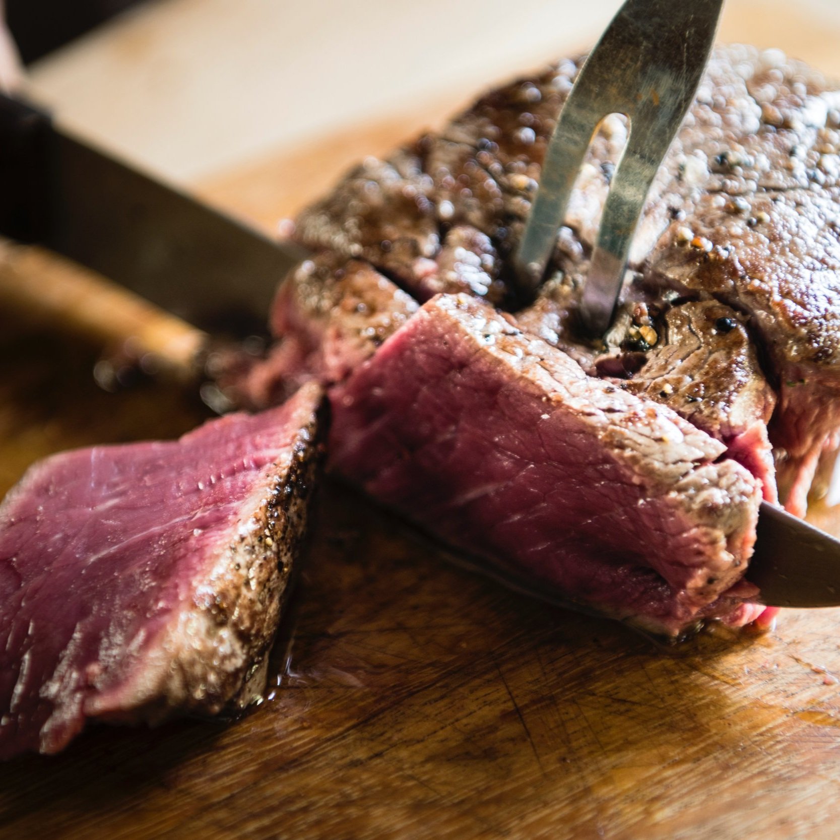 beef_steak_cooked_sliced Juicy, medium-rare steak being sliced on a wooden cutting board, showcasing its tender pink interior and seared crust, with a carving knife and fork positioned for serving.