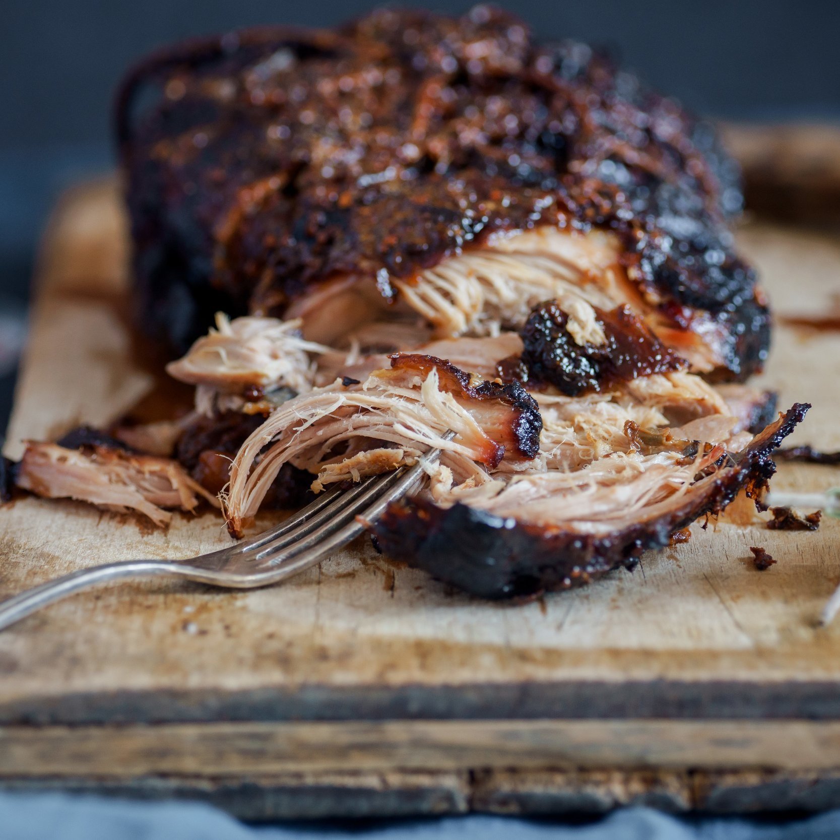 pork_shoulder_bbqorbraised-1 Juicy pulled pork on a wooden cutting board, showcasing tender, shredded meat with a crispy, caramelized exterior. A fork rests beside the meat, emphasizing its delicious texture and flavor.